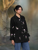 Woman wearing a black floral embroidered shirt and blue jeans standing against a textured gray background.