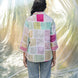 Person wearing a colorful patchwork shirt and jeans standing in front of a textured white fabric.