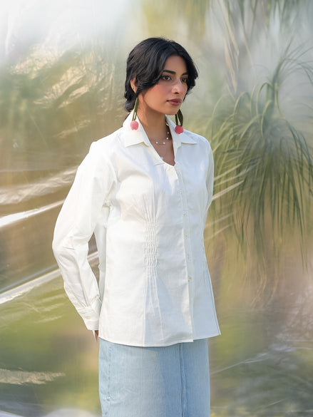 Woman wearing a white shirt and light blue skirt standing in a natural setting with greenery.