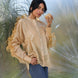 Woman in a beige silk chanderi frilled sleeves shirt and blue jeans holding a glass against a blurred natural background