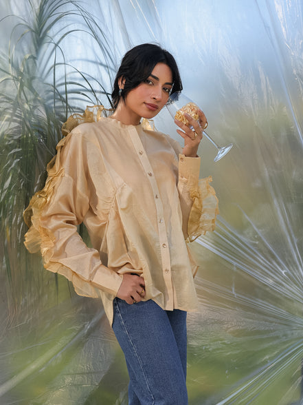 Woman in a beige silk chanderi frilled sleeves shirt and blue jeans holding a glass against a blurred natural background