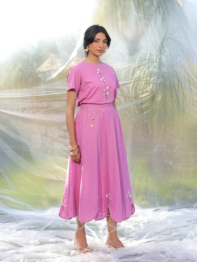 Woman wearing a pink top and skirt with floral details against a blurred natural background
