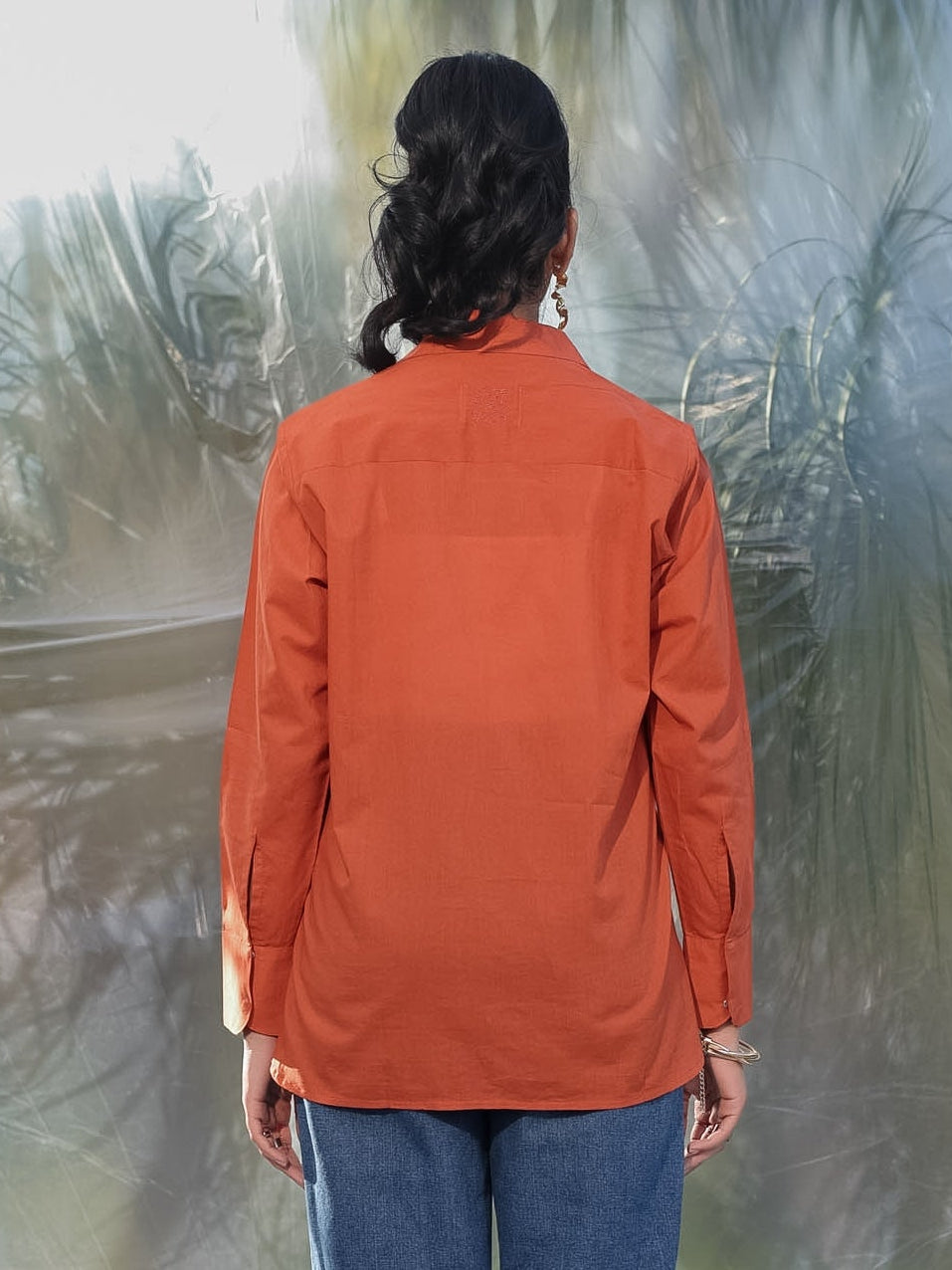 Person wearing a rust shirt and blue jeans standing in front of a textured wall.