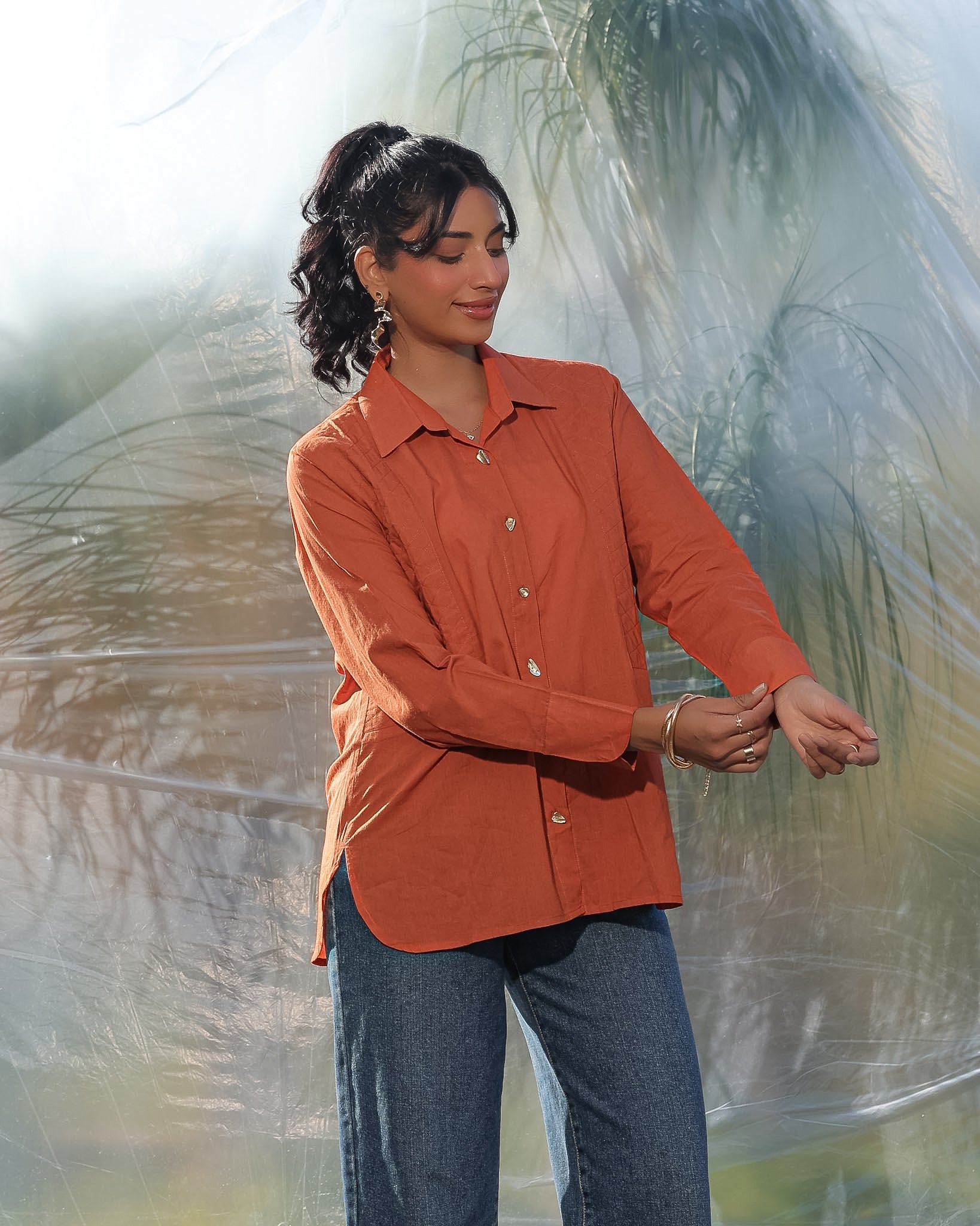 Woman wearing a rust orange shirt and blue jeans standing against a textured white background