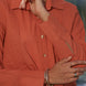 Person wearing a rust-colored shirt with gold buttons with quilted patches in the front, against a blurred natural background.