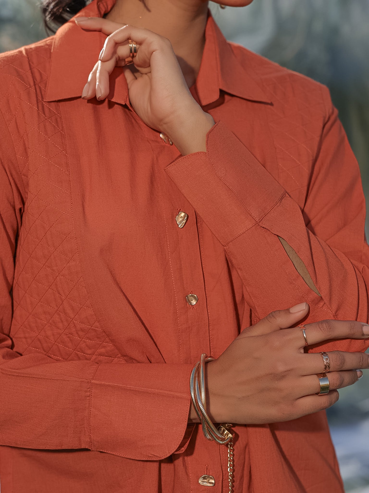 Person wearing a rust-colored shirt with gold buttons with quilted patches in the front, against a blurred natural background.