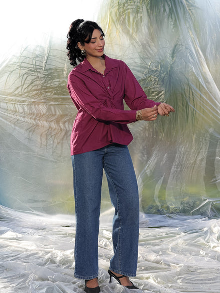 Woman wearing a maroonish wine shirt and blue jeans standing in front of a plastic sheet.