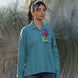 Person wearing a teal shirt with a red flower and a yellow tag, standing on a textured surface.