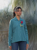 Person wearing a teal shirt with a red flower and a yellow tag, standing on a textured surface.
