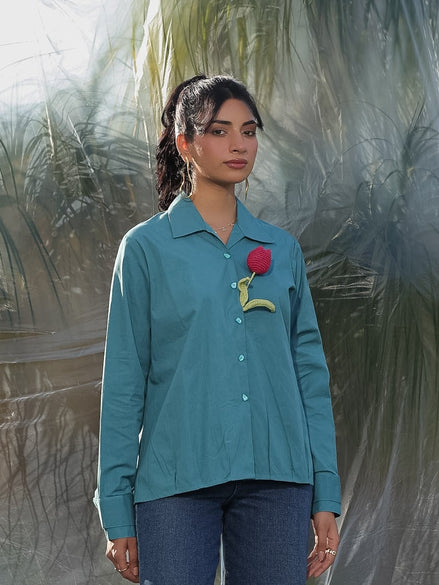 Person wearing a teal shirt with a red flower and a yellow tag, standing on a textured surface.