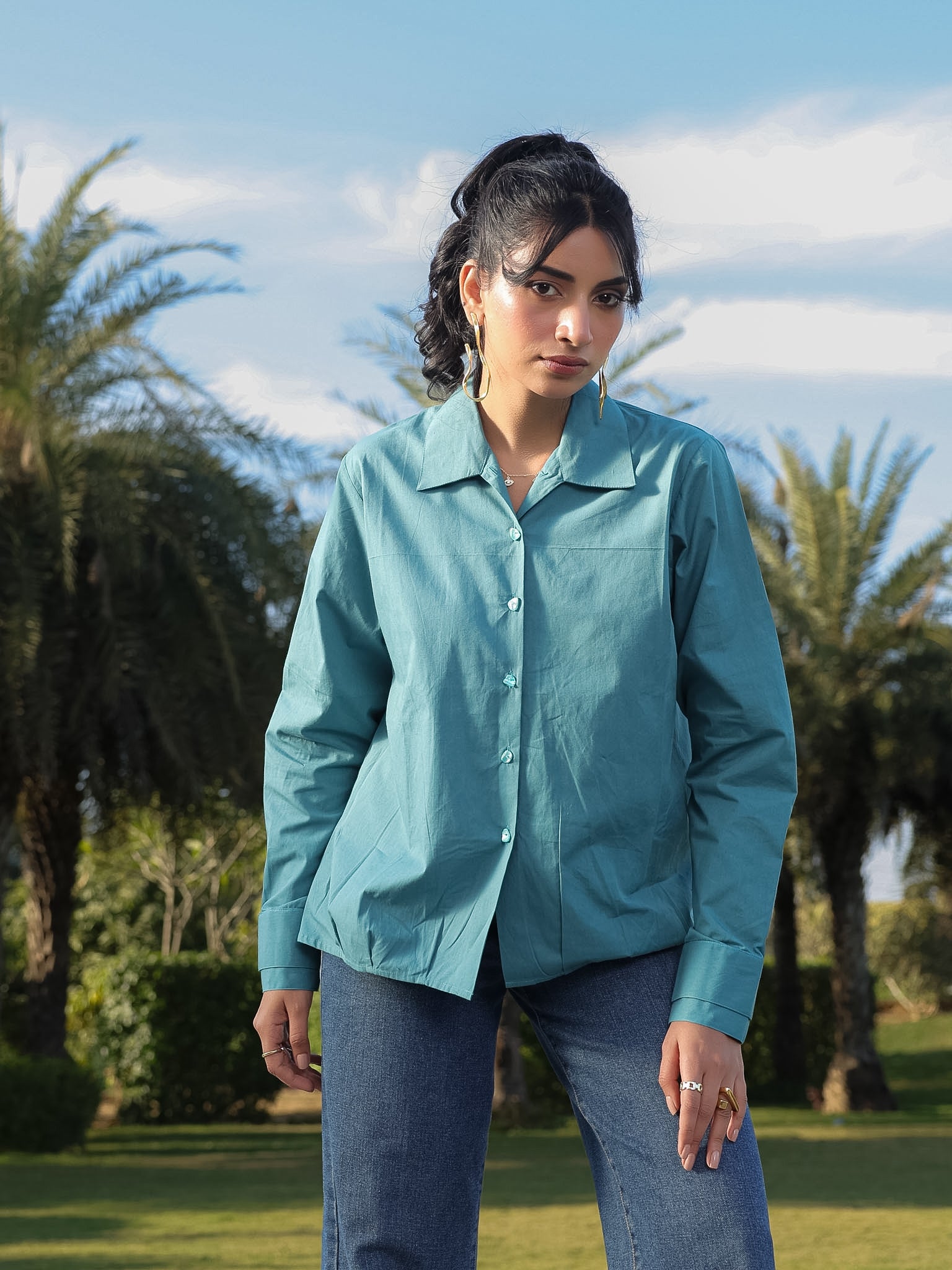 Woman wearing a teal shirt standing outdoors with palm trees in the background