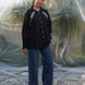 Person wearing a black shirt with white embroidery and blue jeans standing
