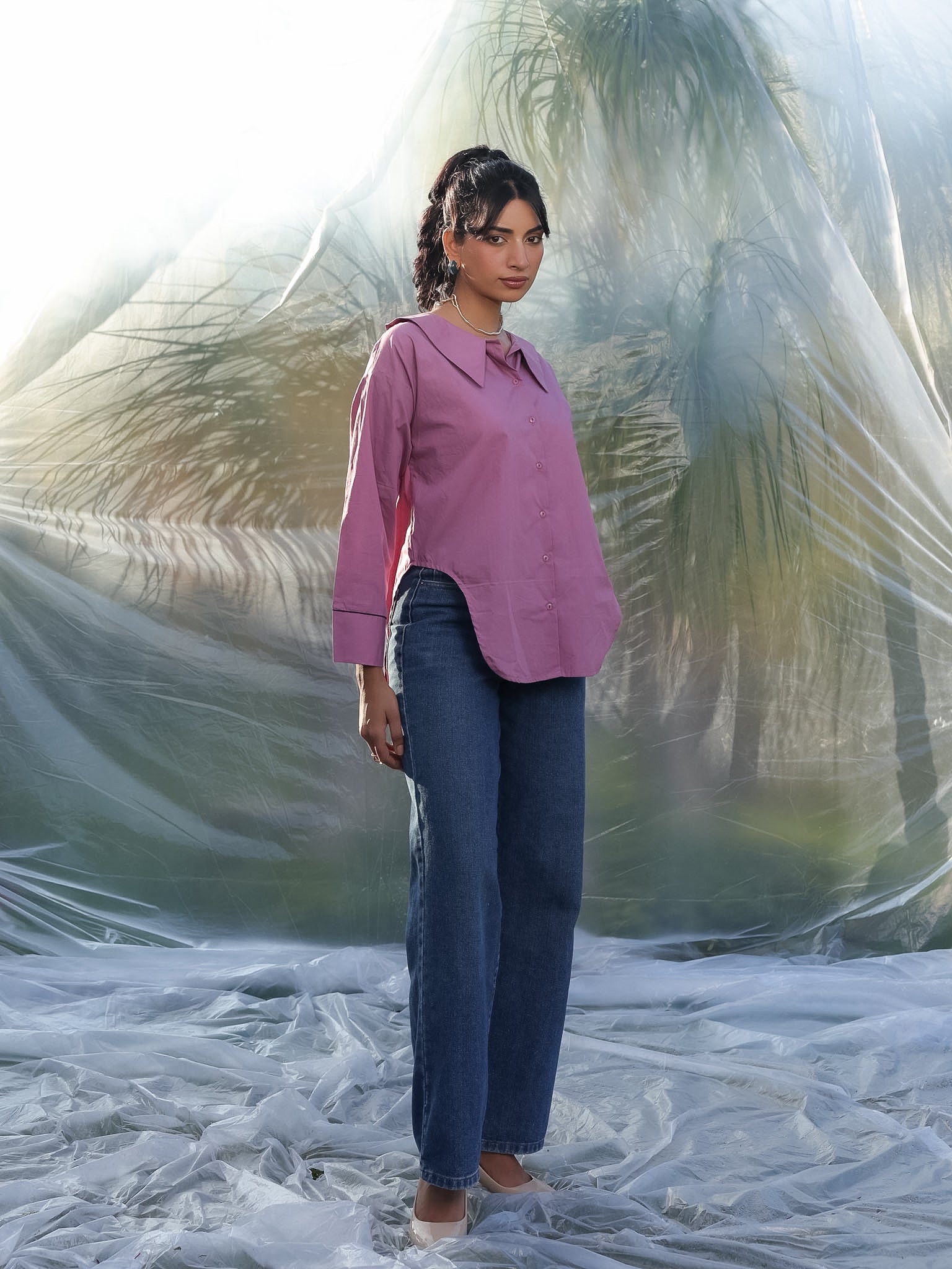 Woman in a mauve pink shirt and blue jeans standing on a beach with palm trees in the background