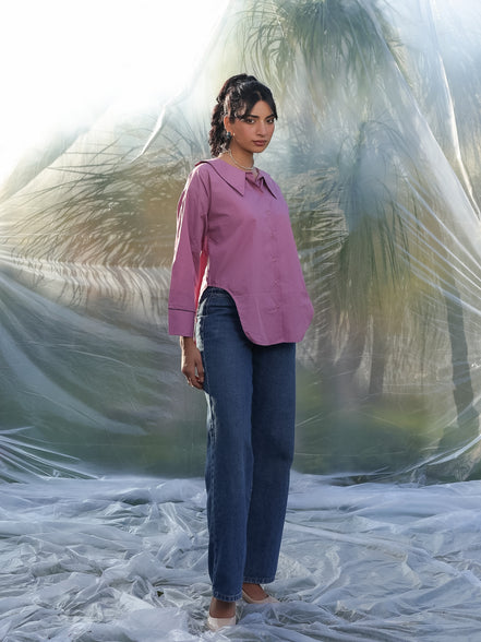 Woman in a mauve pink shirt and blue jeans standing on a beach with palm trees in the background
