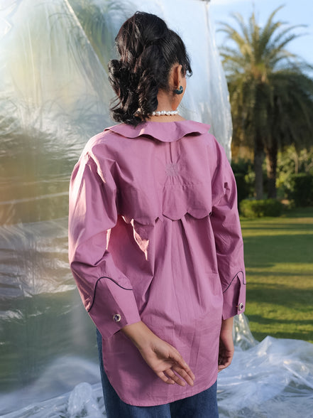 Person wearing a pink shirt with scallop yoke in back in an outdoor setting with plastic sheeting and trees in the background