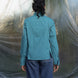 Person wearing a teal blue shirt and blue jeans standing against a textured white wall.