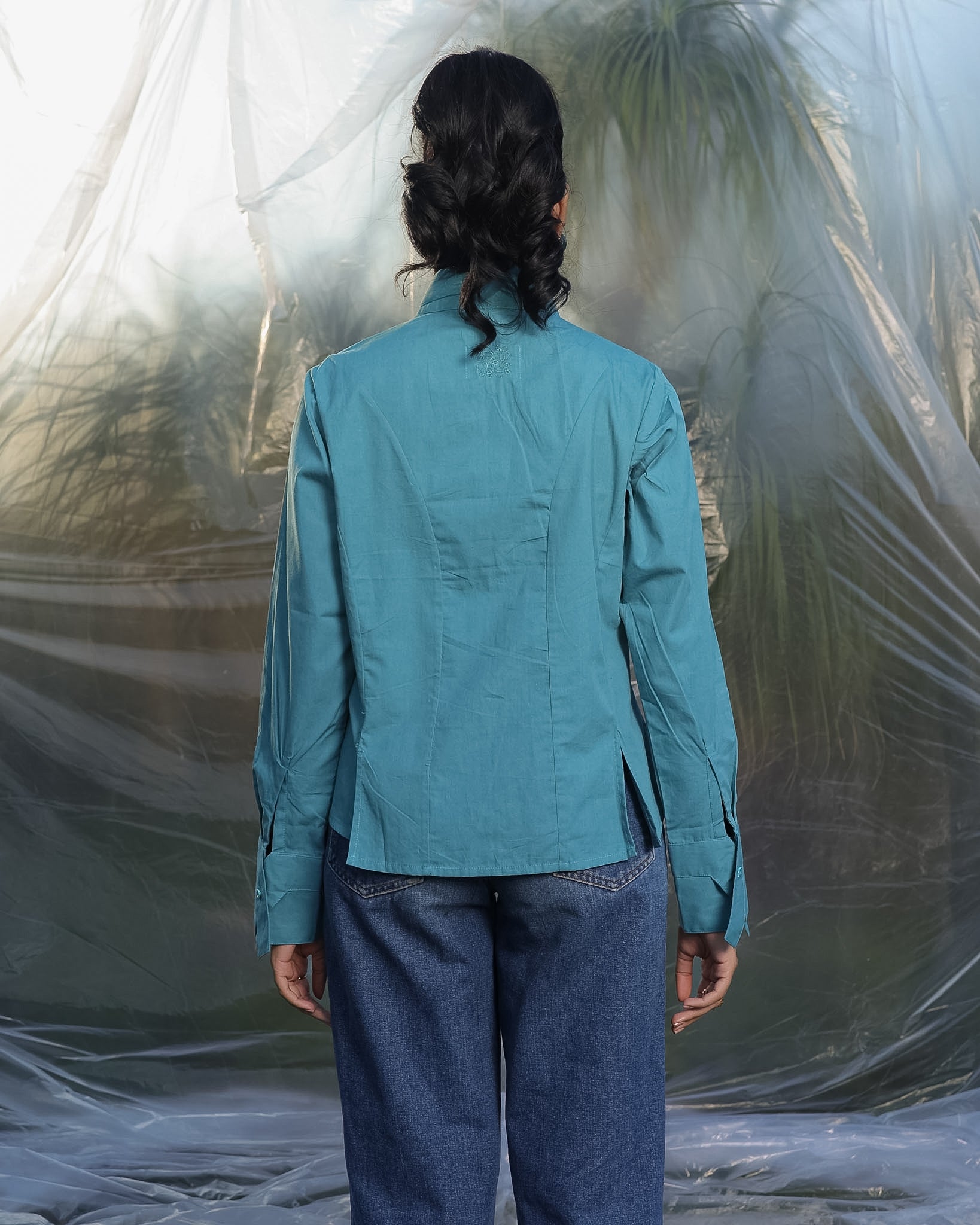 Person wearing a teal blue shirt and blue jeans standing against a textured white wall.