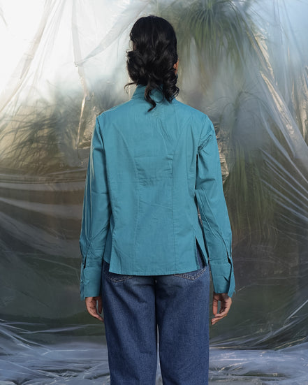 Person wearing a teal blue shirt and blue jeans standing against a textured white wall.
