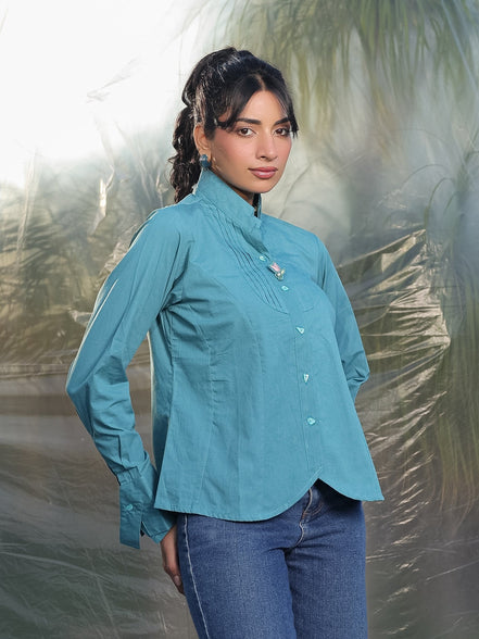 Woman wearing a teal blue shirt and blue jeans standing against a blurred natural background