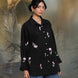 Woman wearing a black floral embroidered shirt and blue jeans standing against a textured gray background.