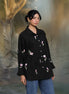 Woman wearing a black floral embroidered shirt and blue jeans standing against a textured gray background.