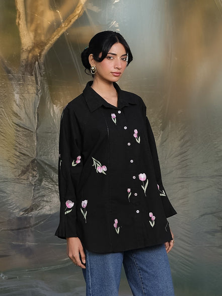 Woman wearing a black floral embroidered shirt and blue jeans standing against a textured gray background.
