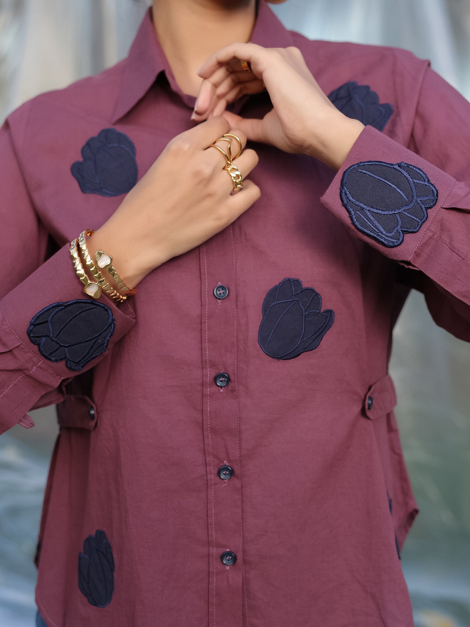 Person wearing a plum shirt with navy blue patches, accessorized with gold jewelry.