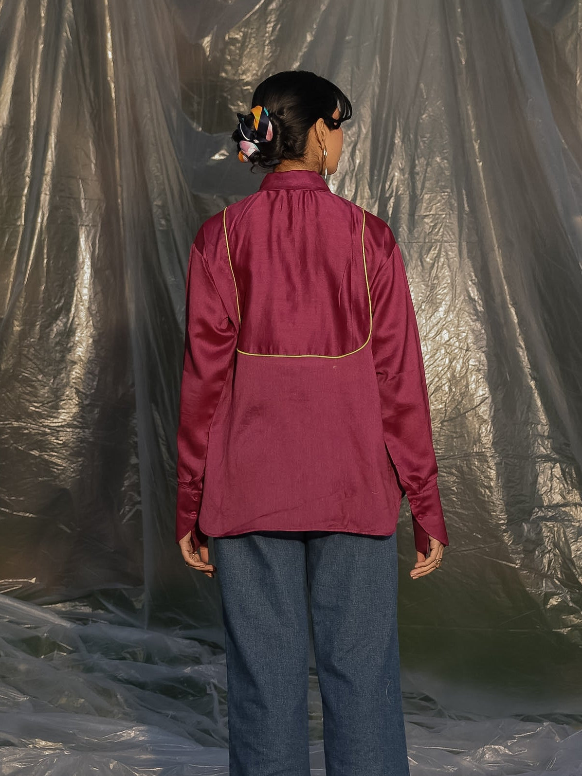 Person wearing a wine silk chanderi shirt and blue pants standing in front of a reflective silver curtain.