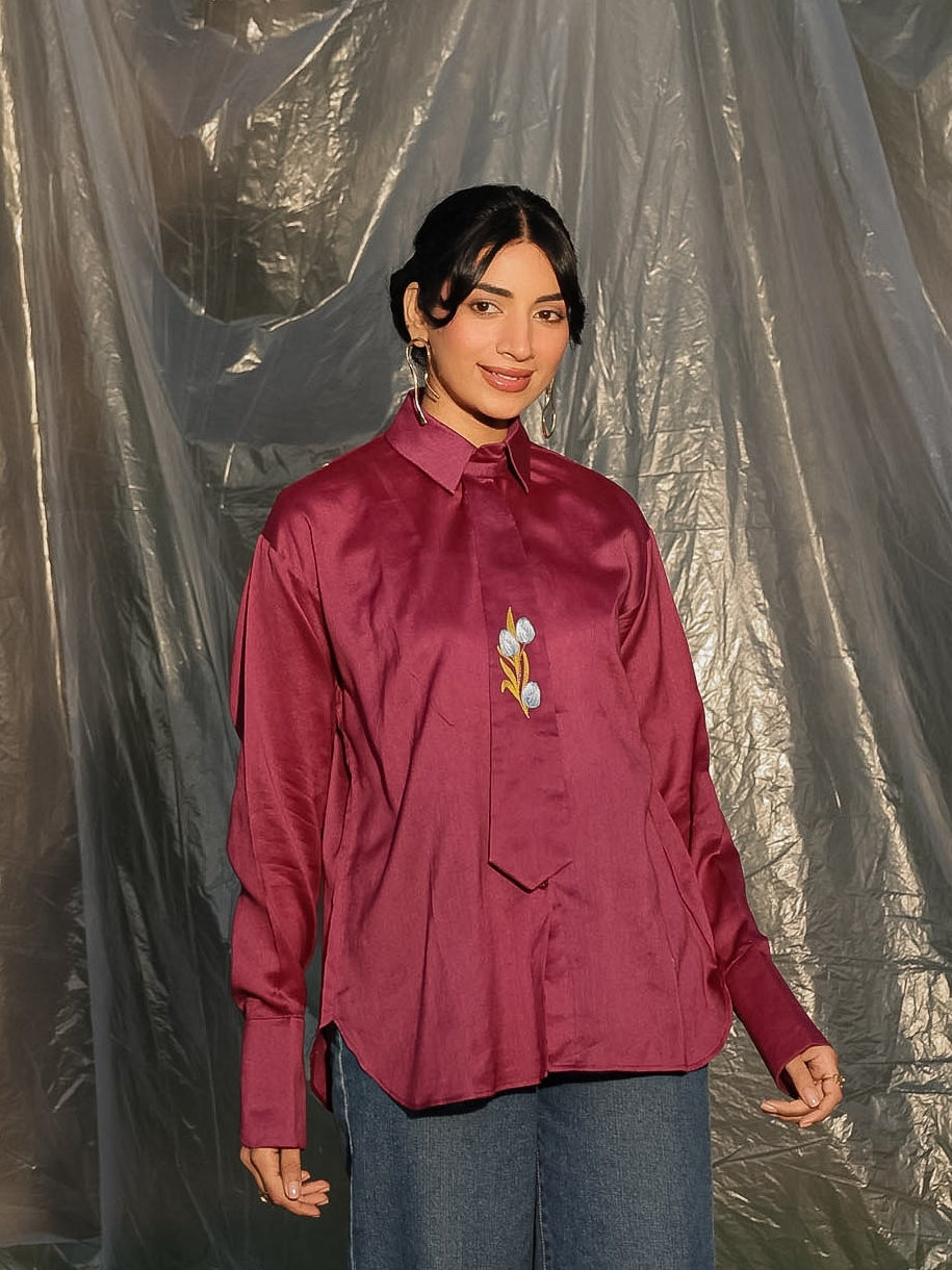 Person wearing a maroon shirt with a tie and blue jeans standing against a tarp background