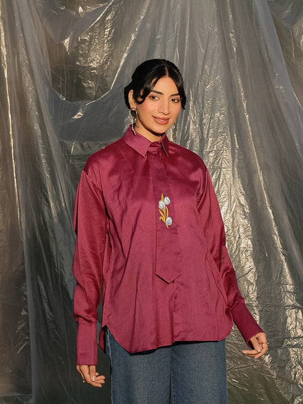 Person wearing a maroon shirt with a tie and blue jeans standing against a tarp background