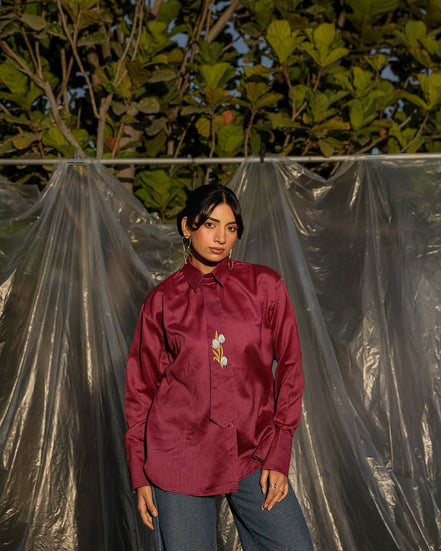 Person wearing a maroon wine silk chanderi shirt with a designer tie, standing against a backdrop of green foliage and plastic sheeting.