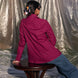 Person sitting on a stool wearing a winish maroonish shirt and blue jeans, with a silver tarp in the background.