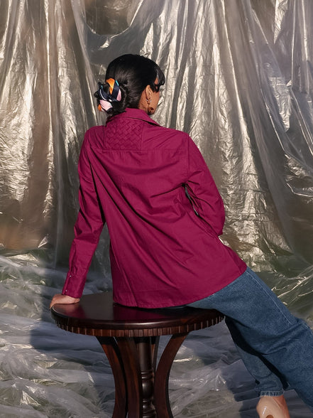 Person sitting on a stool wearing a winish maroonish shirt and blue jeans, with a silver tarp in the background.