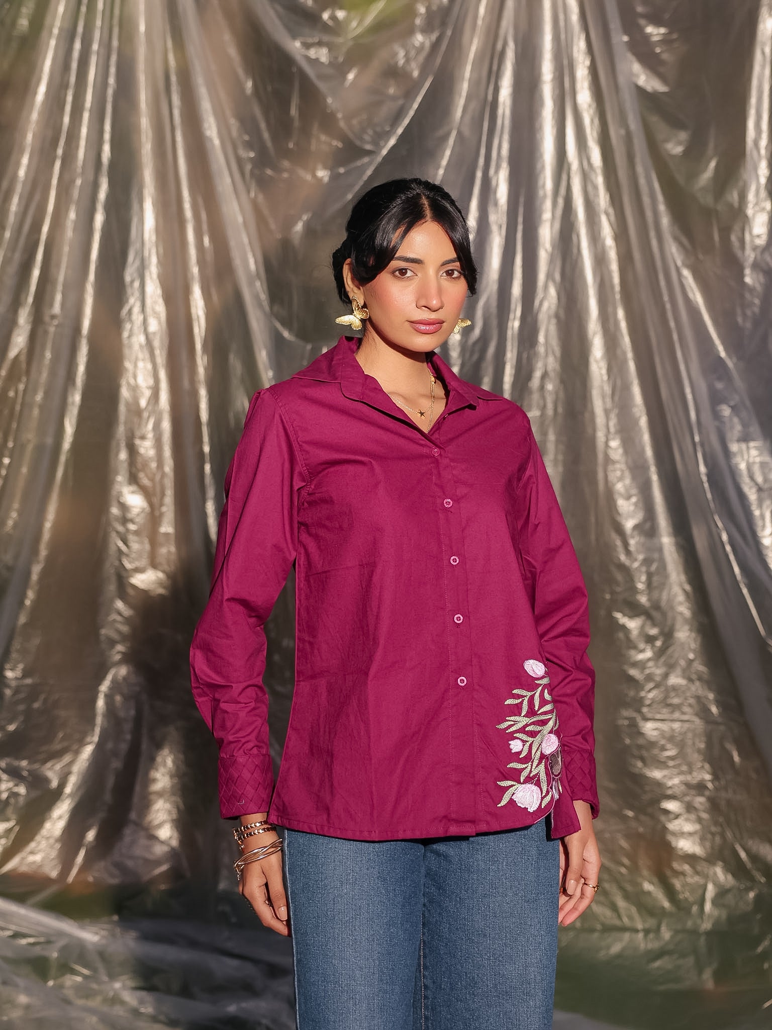 Woman wearing a magenta shirt with floral embroidery against a reflective silver curtain.